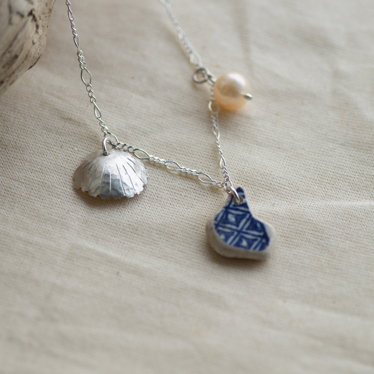 A delicate silver-toned necklace with a textured shell-shaped charm, a small blue-and-white patterned stone charm, and a single pearl bead, displayed on a beige linen fabric background.