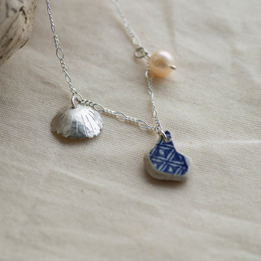 A delicate silver-toned necklace with a textured shell-shaped charm, a small blue-and-white patterned stone charm, and a single pearl bead, displayed on a beige linen fabric background.