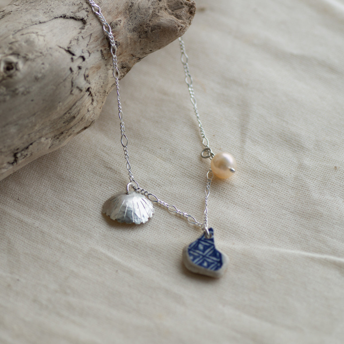 A delicate silver-toned necklace with three charms: a silver seashell, a small white pearl bead, and a blue-and-white patterned ceramic tag, displayed on a beige fabric surface next to a piece of driftwood.