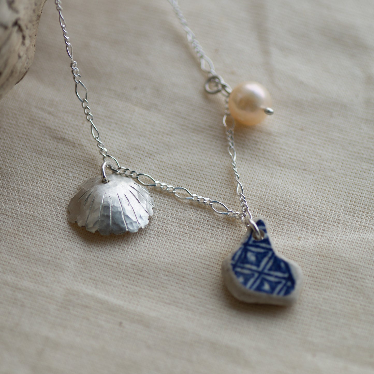 A silver chain necklace with three charms: a textured silver shell, a small blue and white patterned ceramic piece, and a single pearl bead, displayed on a beige linen fabric background.
