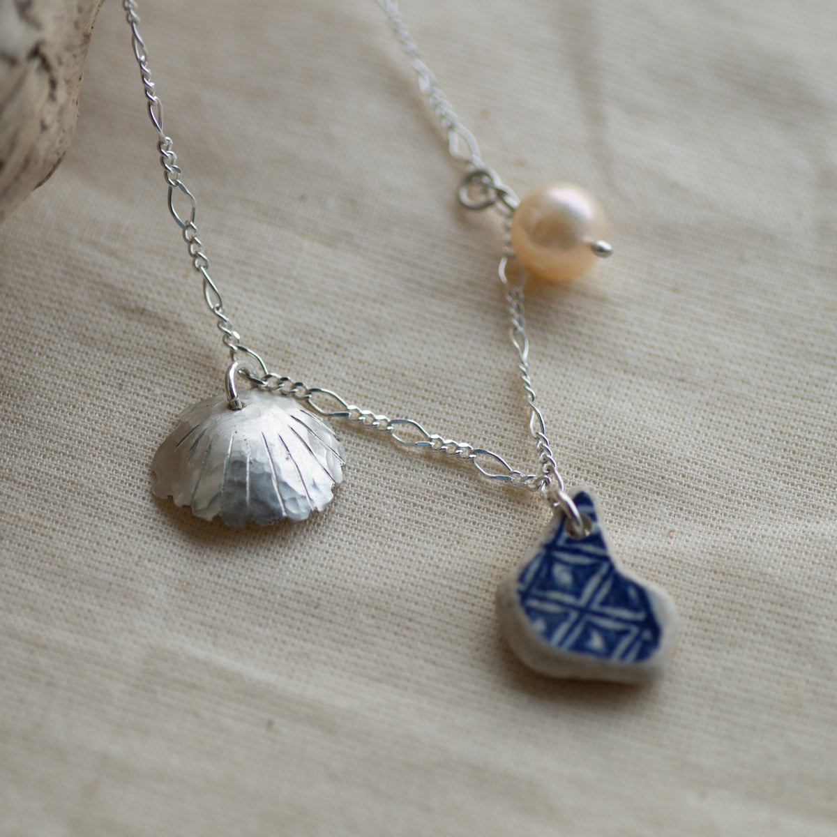 A silver chain necklace with three charms: a textured silver shell, a small blue and white patterned ceramic piece, and a single pearl bead, displayed on a beige linen fabric background.