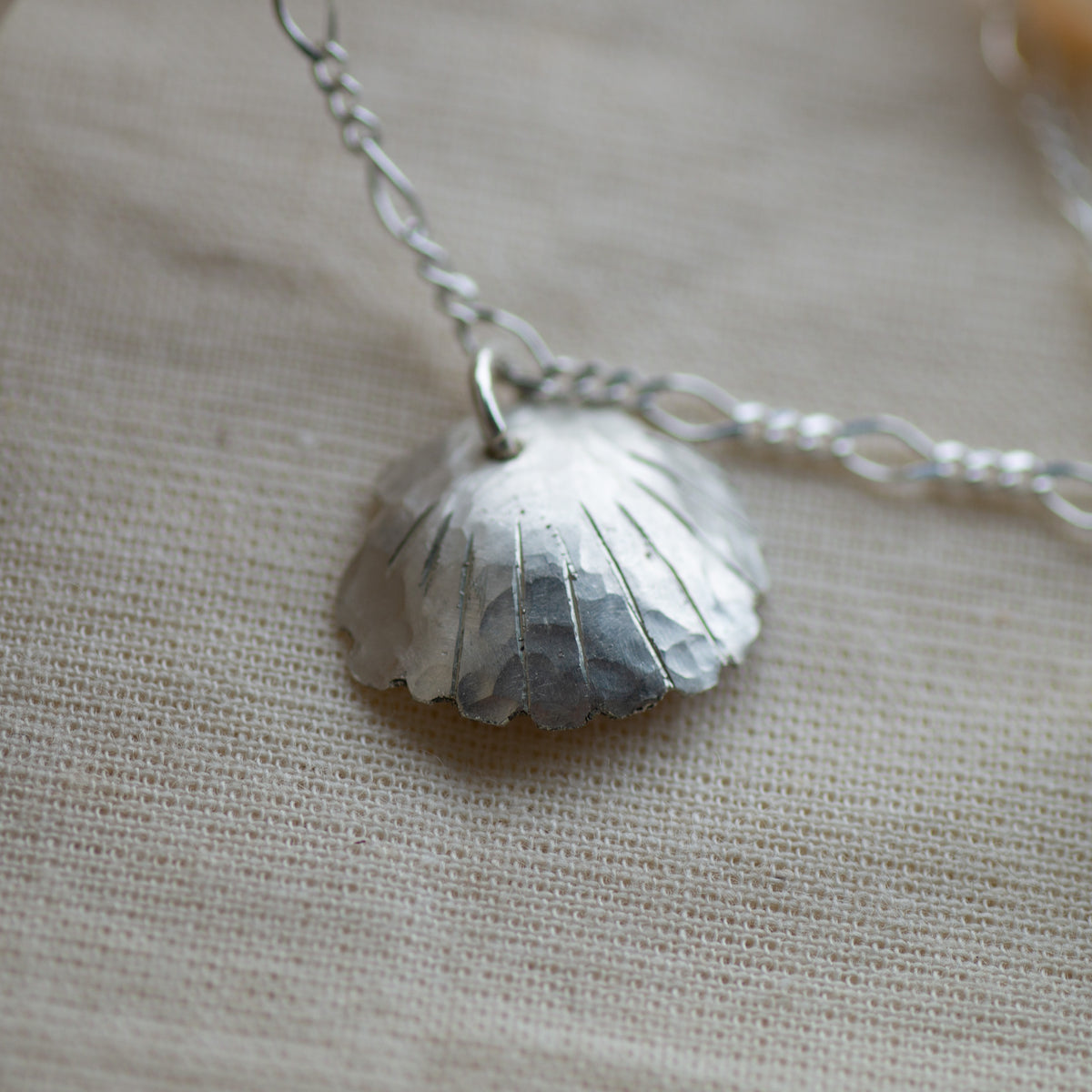 A hammered silver shell pendant necklace on a delicate silver chain, displayed on a textured beige linen fabric background.